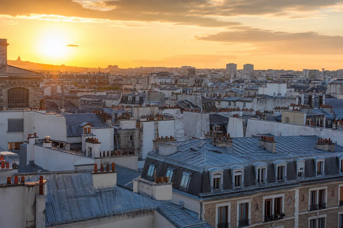 Vue en plongée sur des toits en zinc parisiens et des façades d’immeubles anciens du 20e arrondissement au soleil couchant