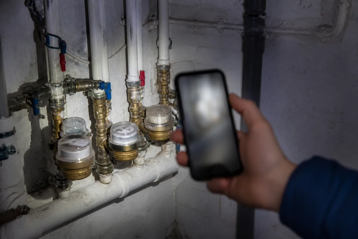 Main tenant un smartphone devant une canalisation apparente dans un local technique d’immeuble